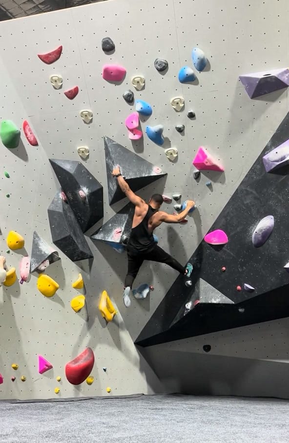 Rohin mid-climb on an indoor bouldering wall, reaching between colourful holds