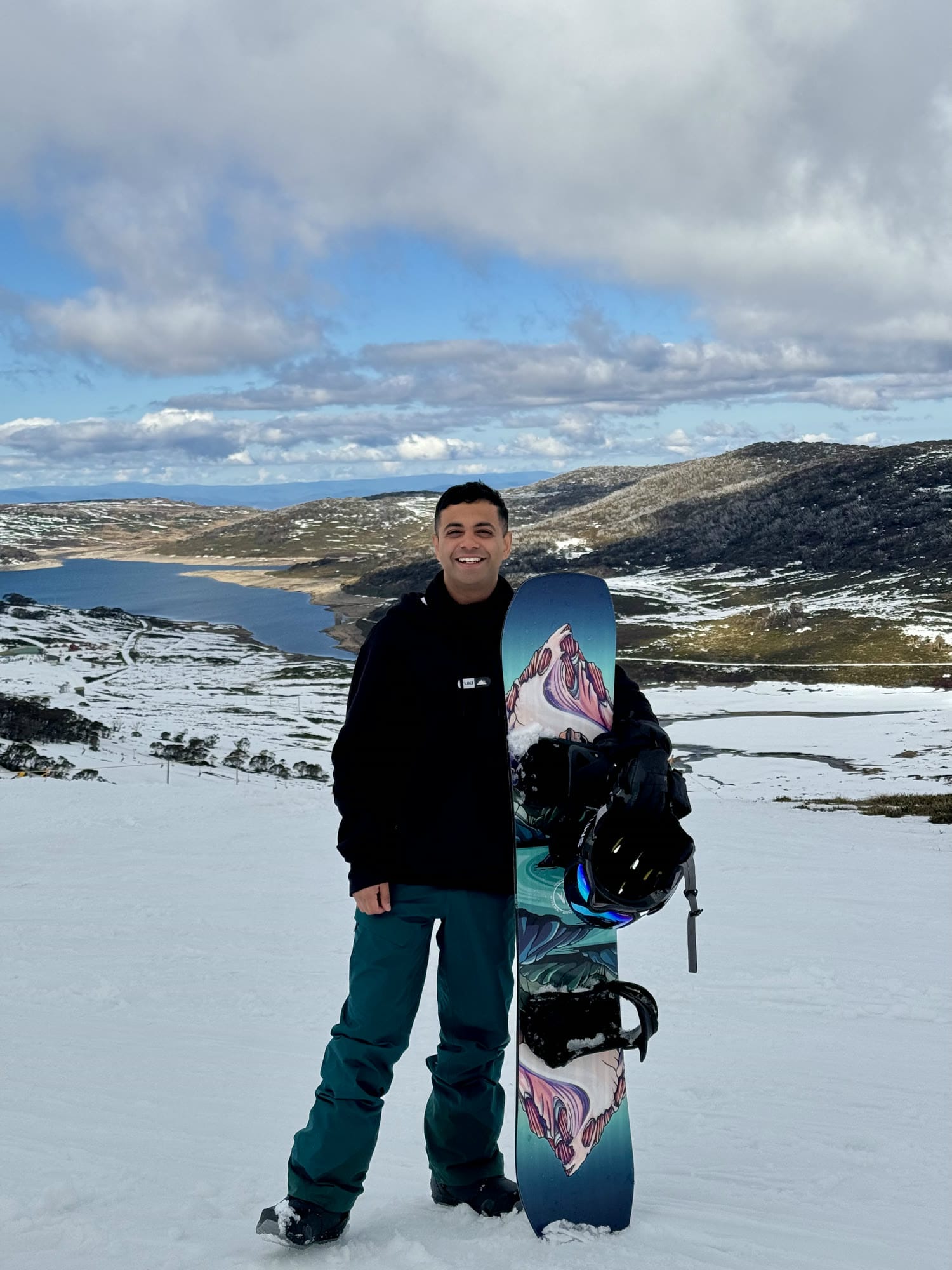 Rohin smiling on a snowy slope, holding his snowboard, with a lake and hills behind him