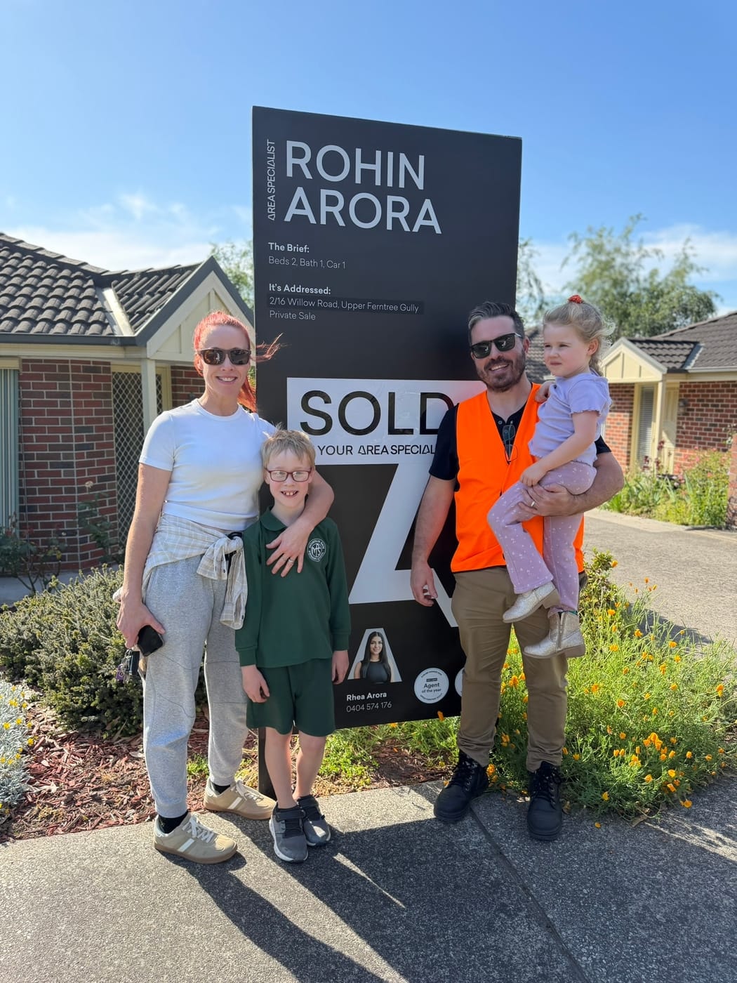 A family on a sunny footpath in front of their SOLD signboard, the father in a hi-vis vest holding a toddler