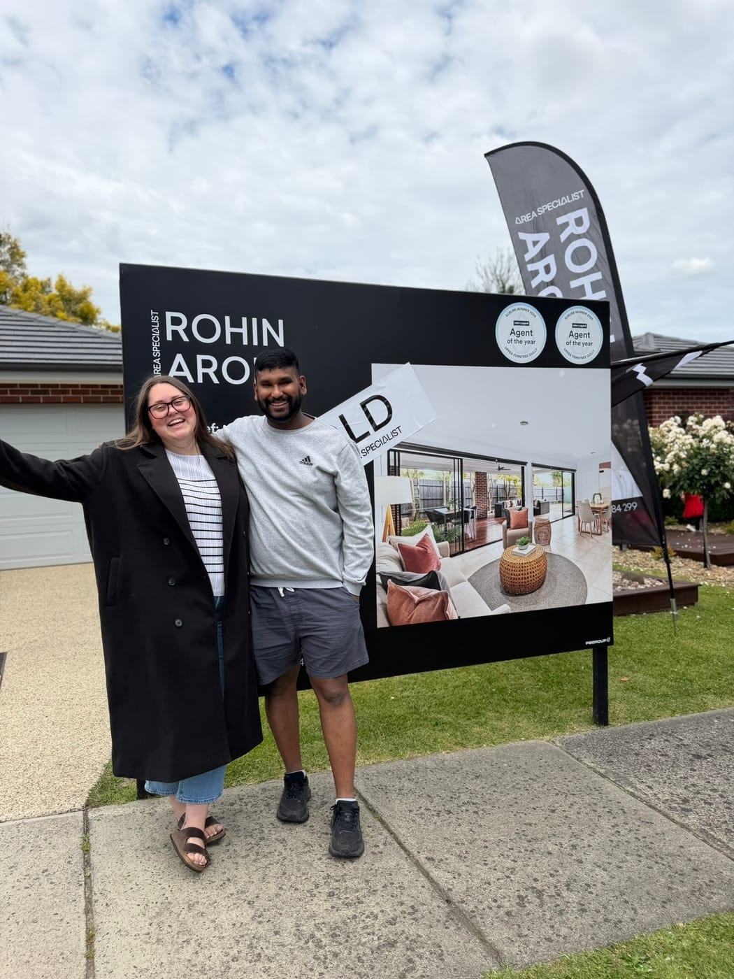 A couple with arms outstretched beside a SOLD signboard and a Rohin Arora flag
