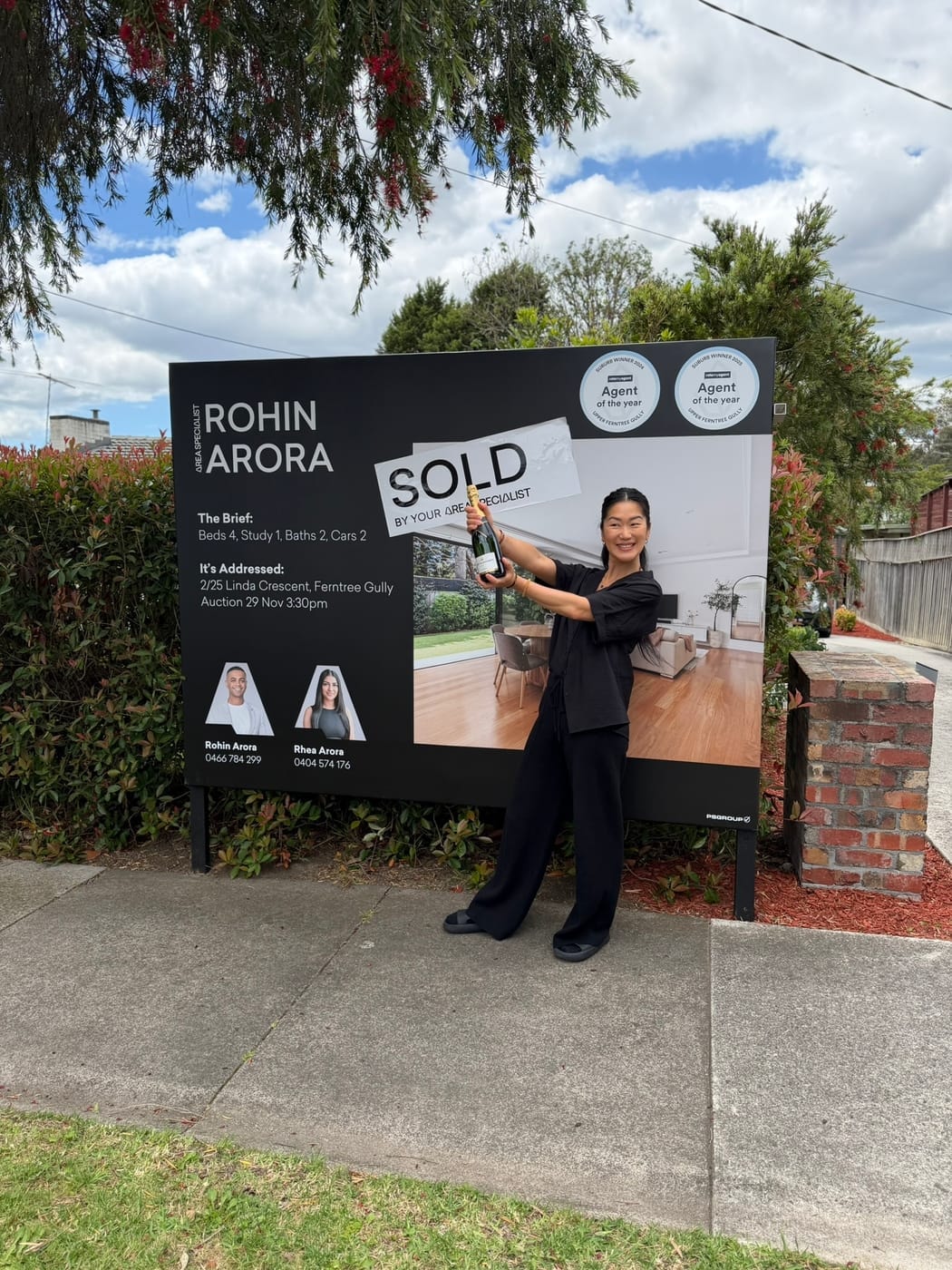 A woman raising a bottle of champagne in front of a SOLD signboard