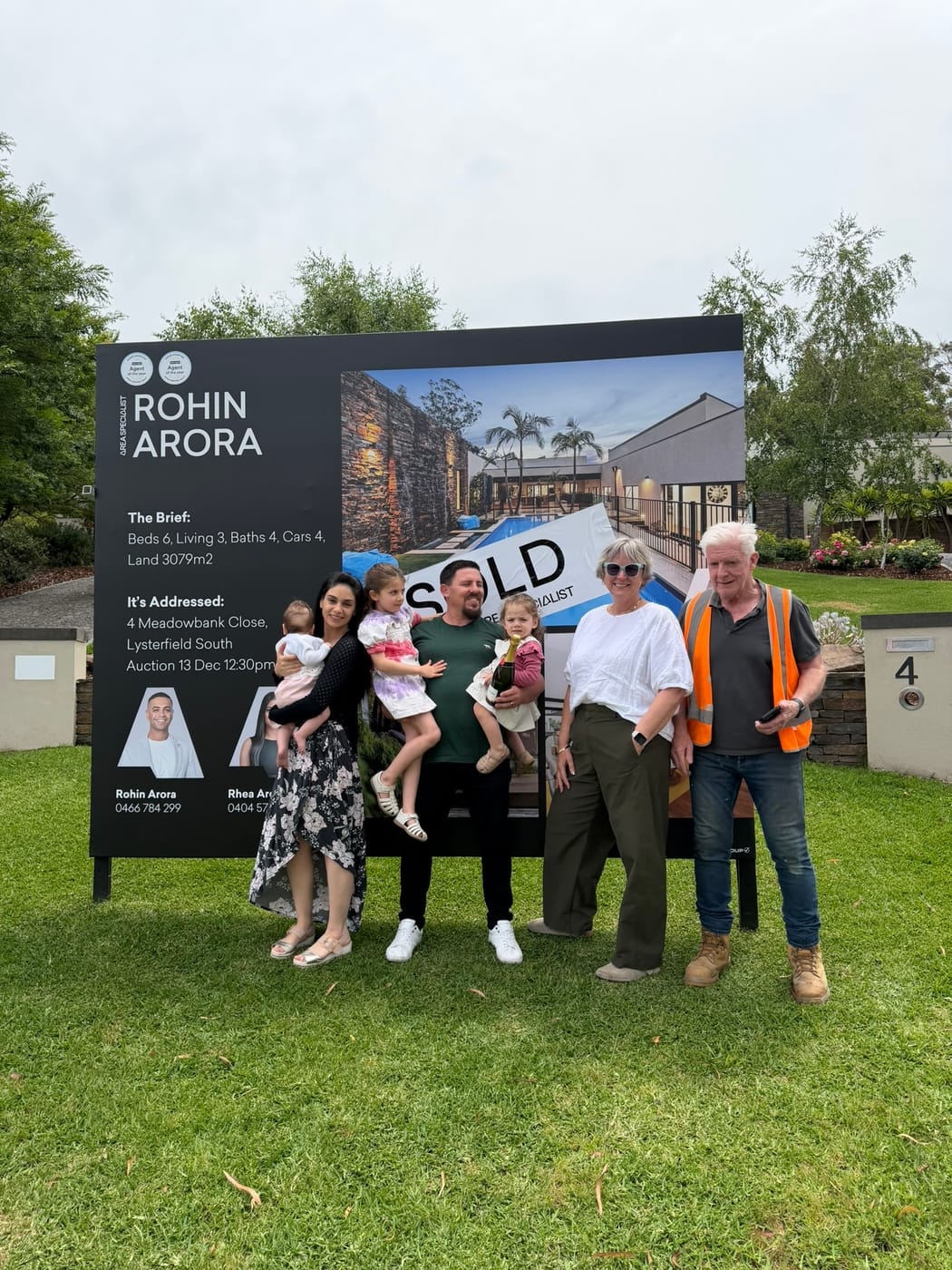 A family with young children and grandparents on the lawn in front of a SOLD signboard