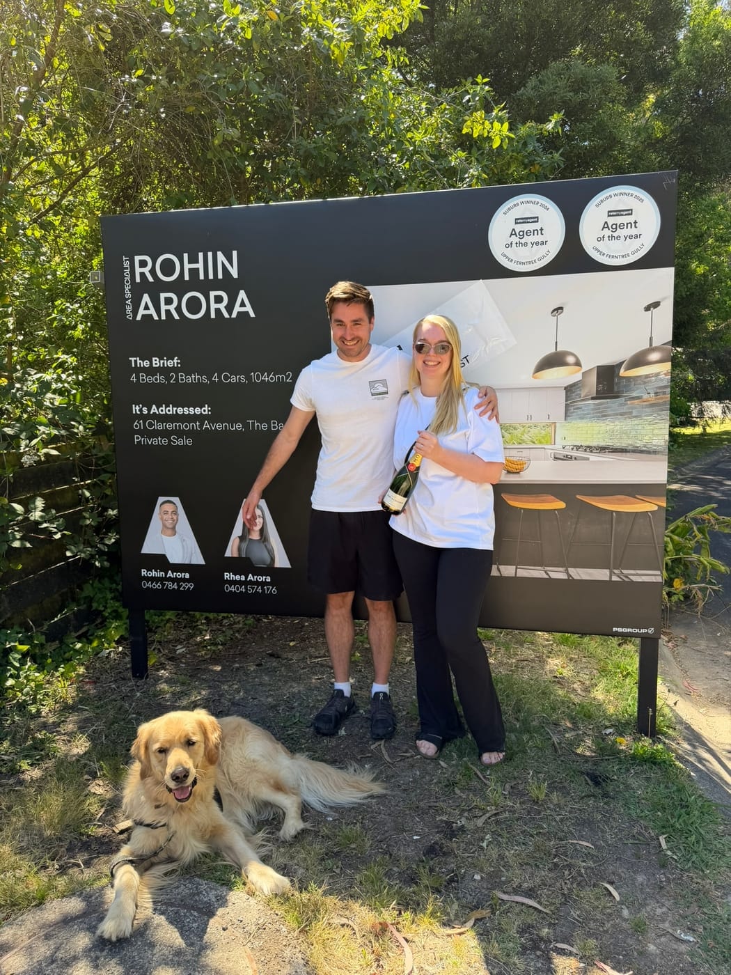 A couple holding champagne beside their SOLD signboard, a golden retriever lying in the grass at their feet