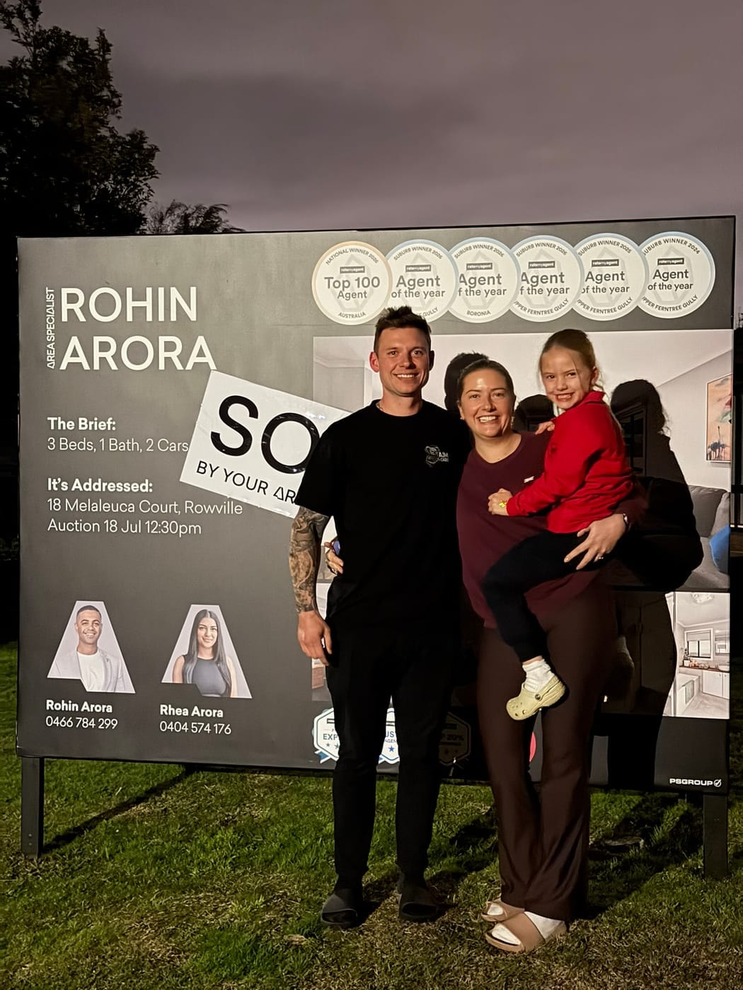 A couple and a girl in a red jumper in front of a SOLD signboard at dusk