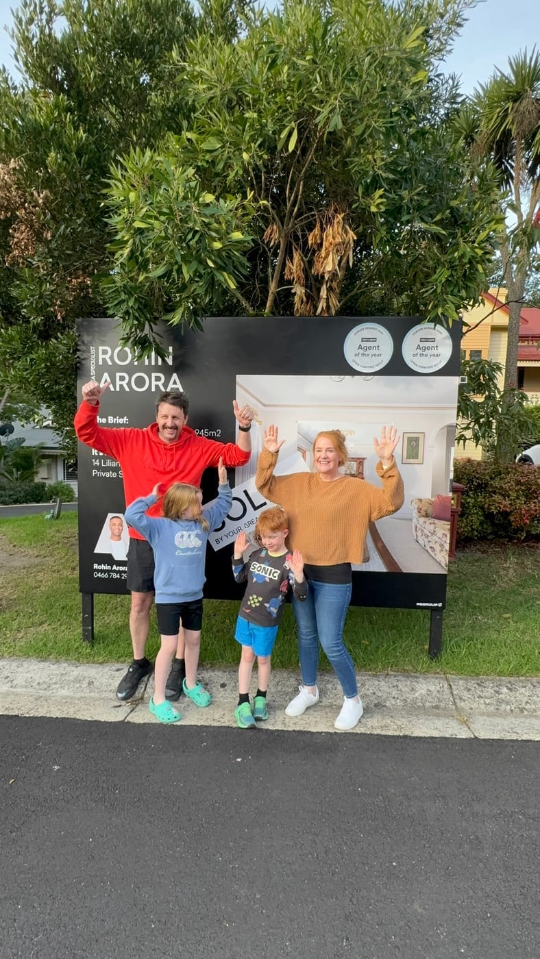 A family of four throwing their arms up in front of a SOLD signboard