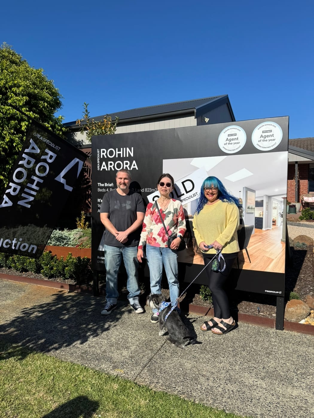 Three people and a small dog on the footpath in front of a SOLD signboard