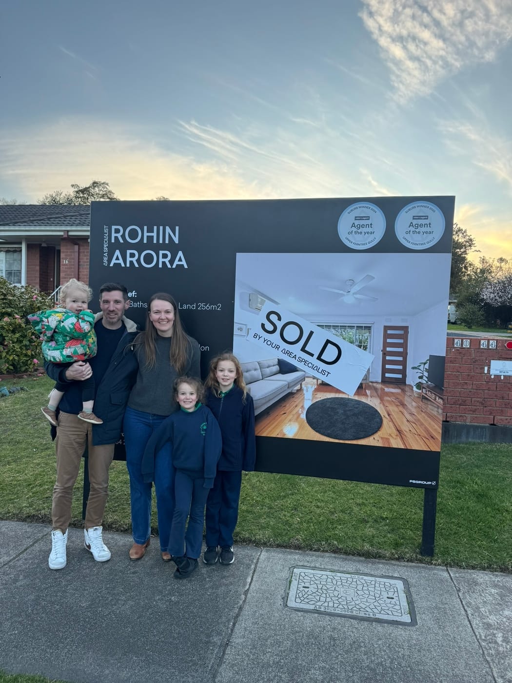A couple and three young children in front of a SOLD signboard in late-afternoon light