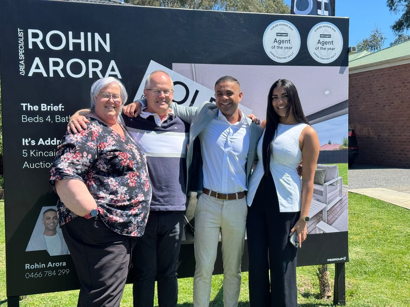 Four people standing close together, smiling, in front of a SOLD signboard
