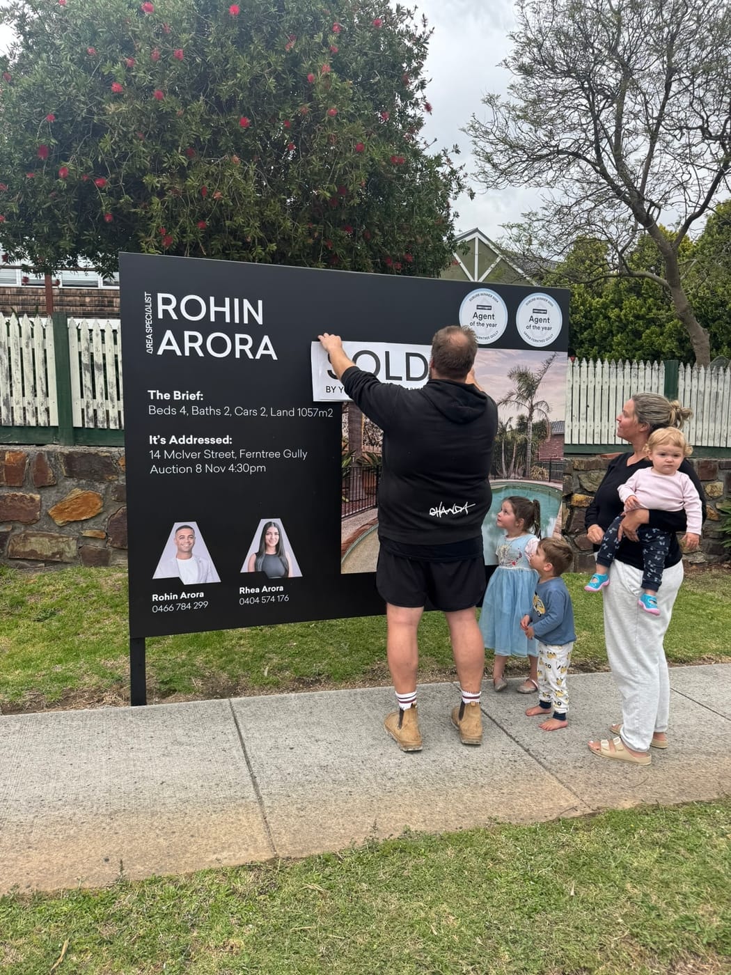 A man pressing the SOLD sticker onto the signboard as his family looks on