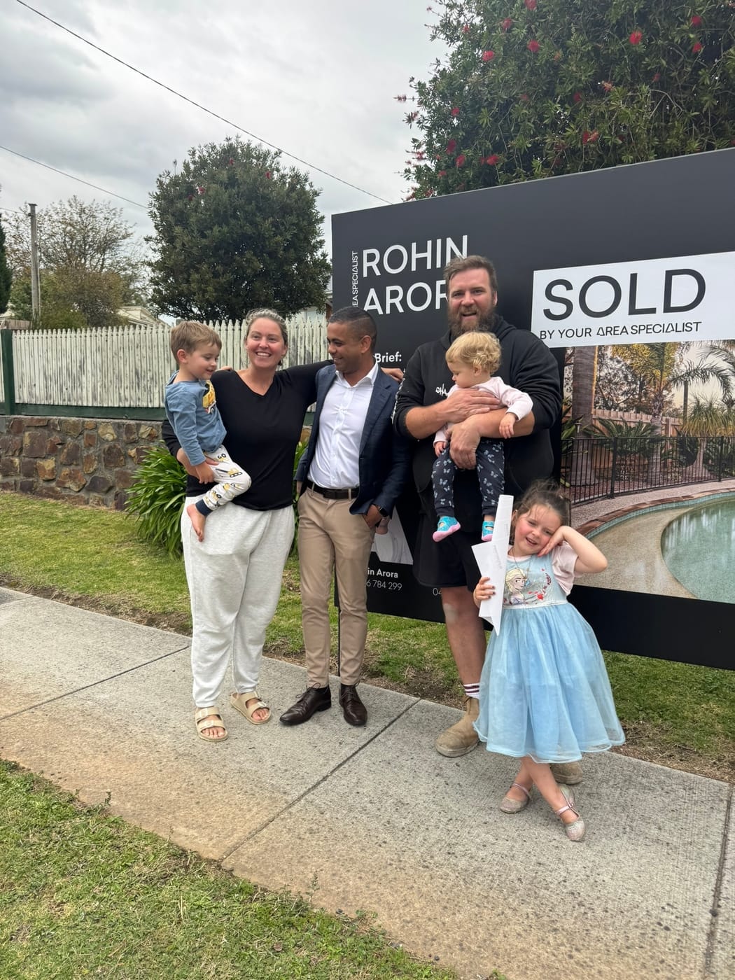 A family with young children, one in a blue party dress, beside a SOLD signboard
