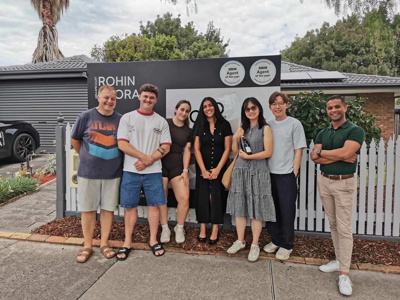 Seven people lined up along a white picket fence in front of a SOLD signboard