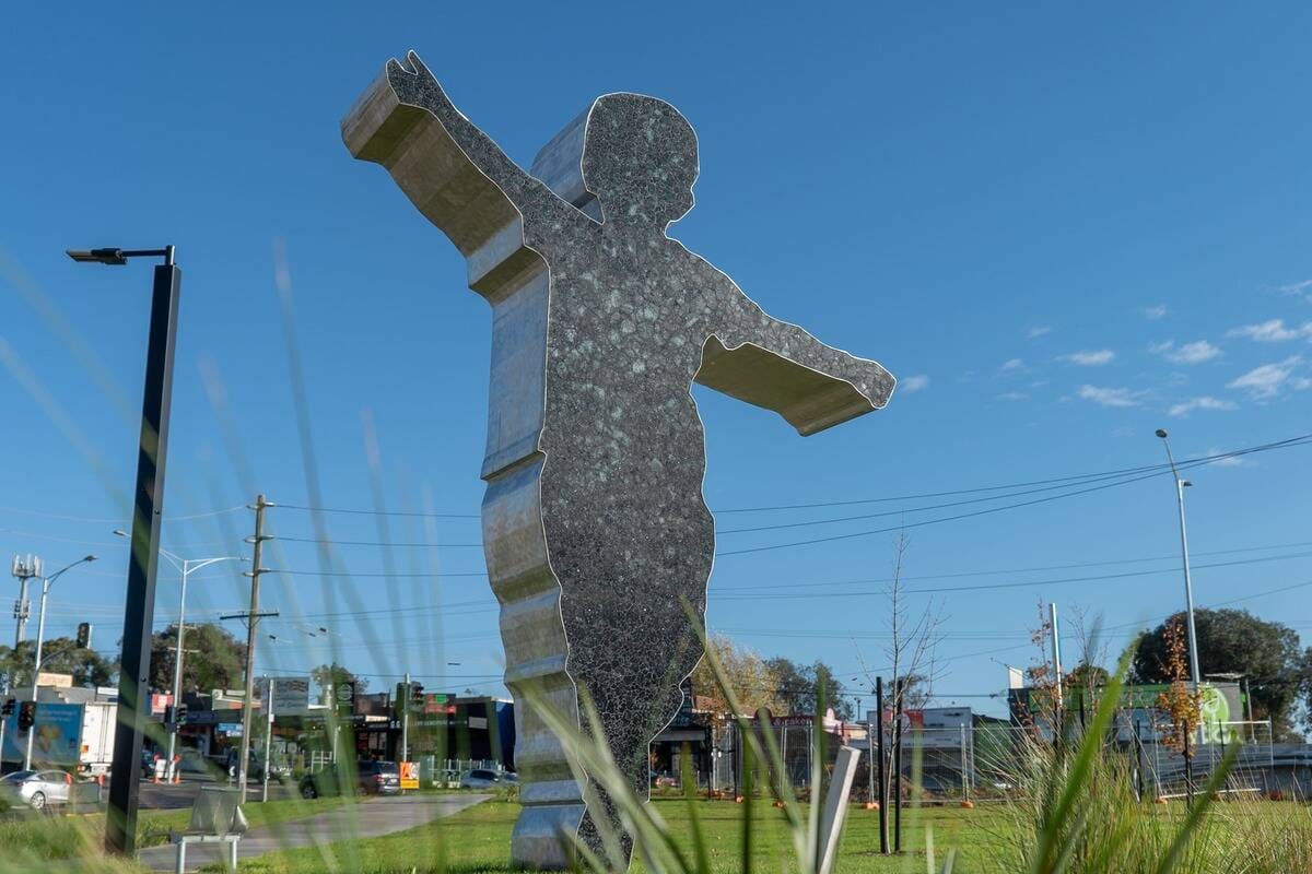 A tall mosaic-clad sculpture of a boy with his arms spread like wings, on a grassy reserve beside a Bayswater road